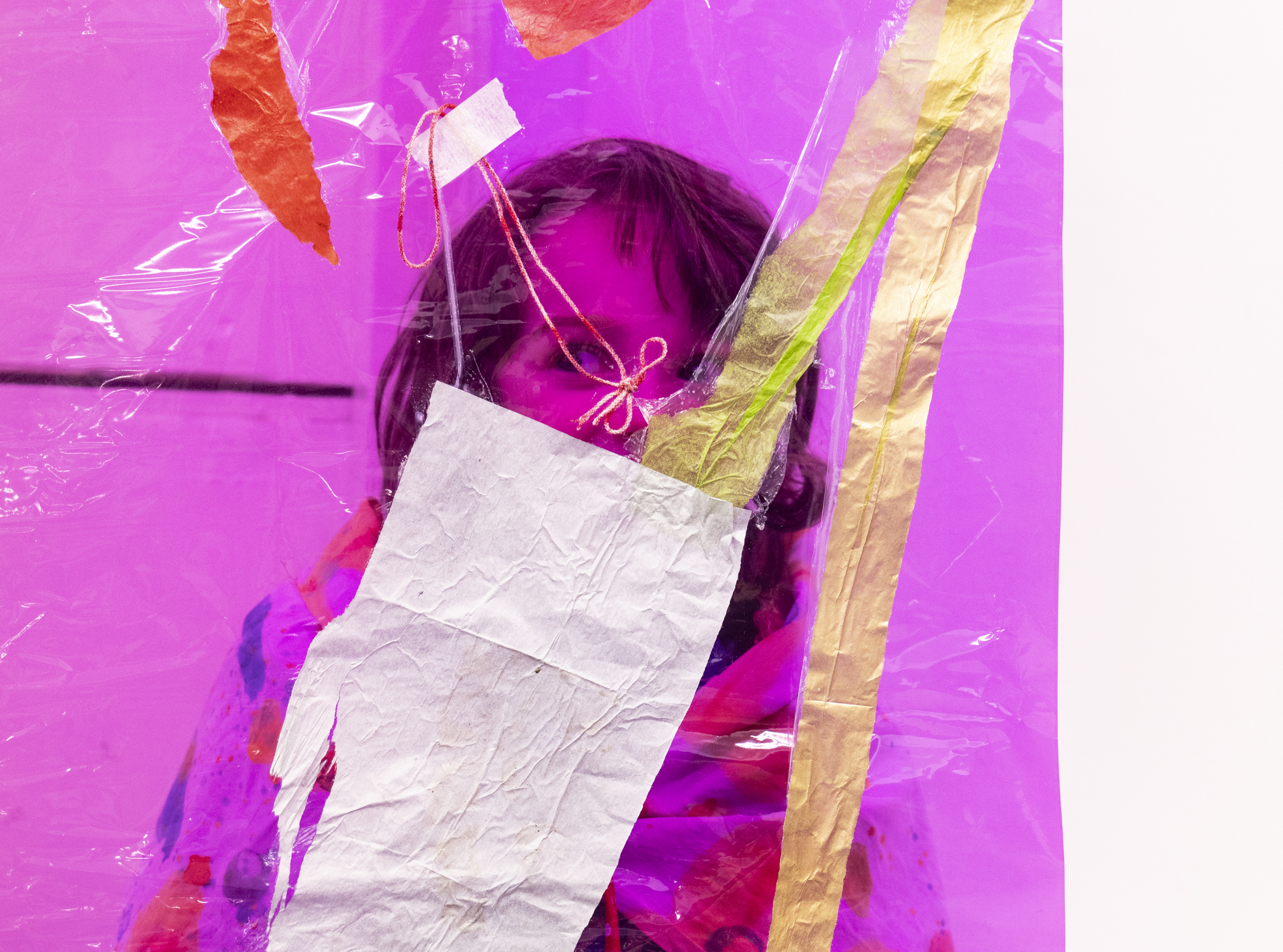 Image description: Closeup of a child's face looking through a large piece of pink cellophane decorated with coloured tape and string, in an art exhibition.