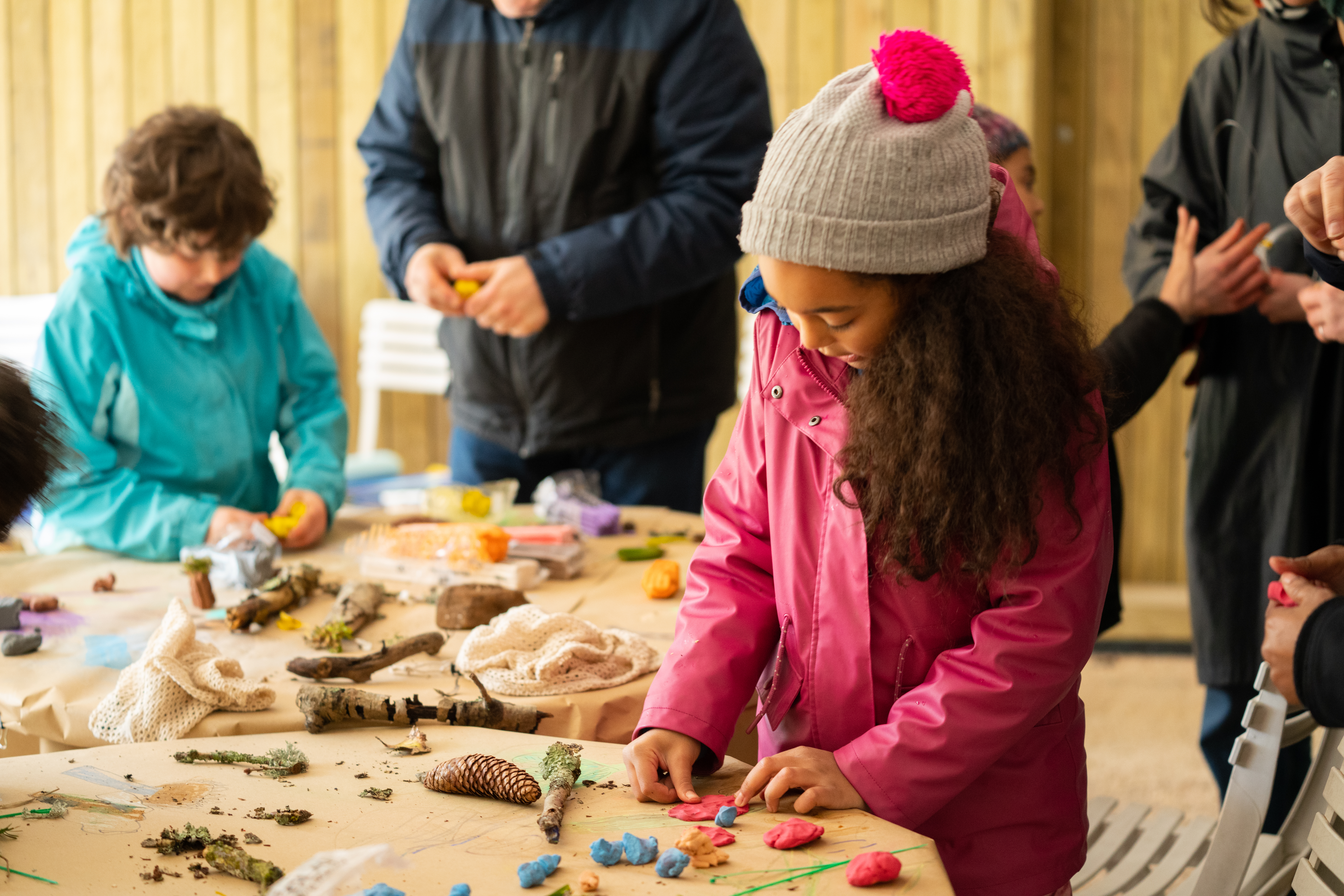 photograph of a child in a pink jacket and hat looking downwards whilst working on plasticine on a table, with other children, in a covered area outdoors.