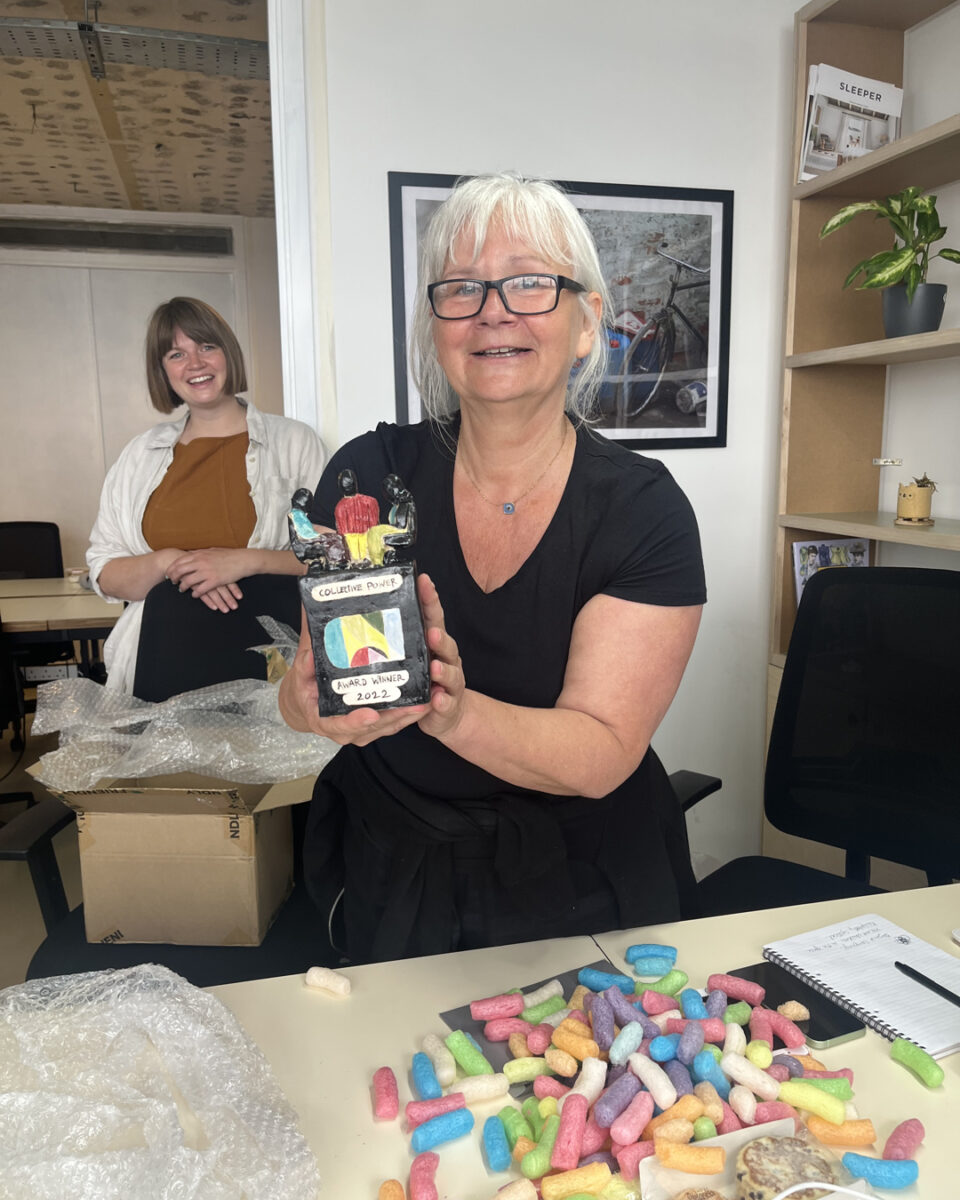 Lisa Nash (ACAVA Curator, Social Practice) unboxes the award in the office with other members of the team. The award is a 20 cm high sculpture in black, yellow, blue and white glazed ceramics of three people sitting and facing each other, above a cubic shape with the award's logo.