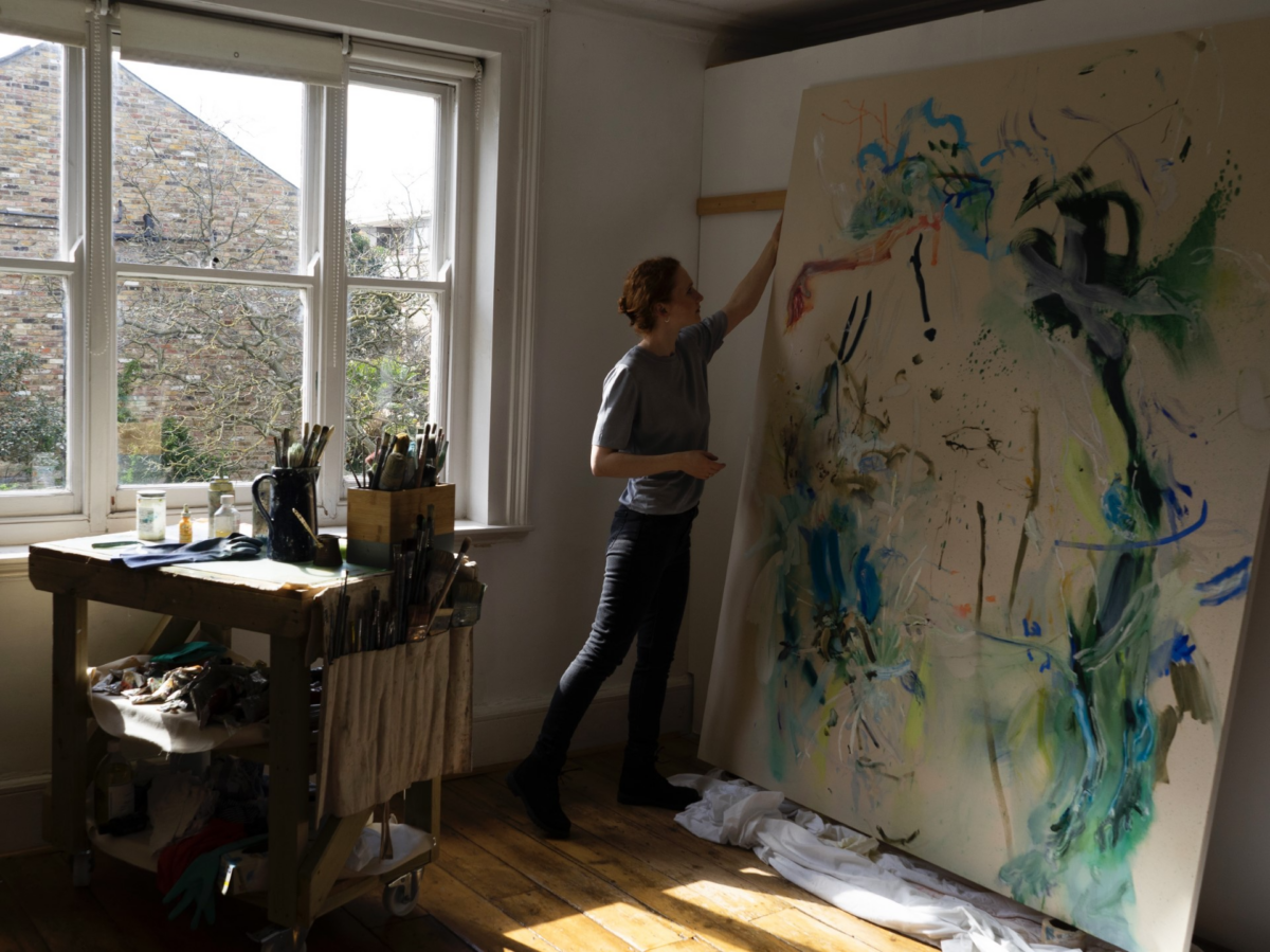Photograph of an artist working on a large painting in a studio bathed in natural light.