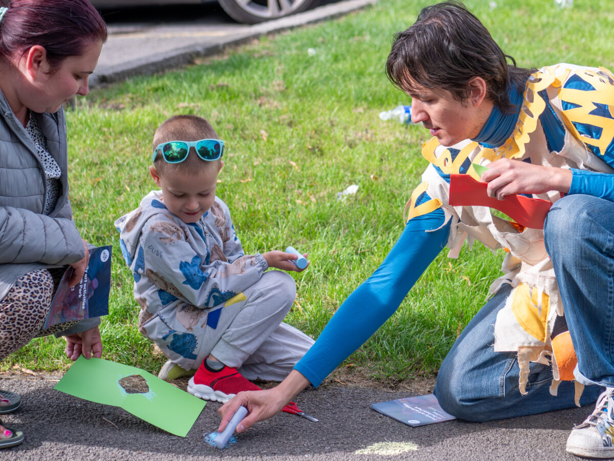 Close-up photograph of ACAVA's Project Assistant Maxi Himpe and a young family using colourful chalk to mark the tarmac, outdoors in a park.