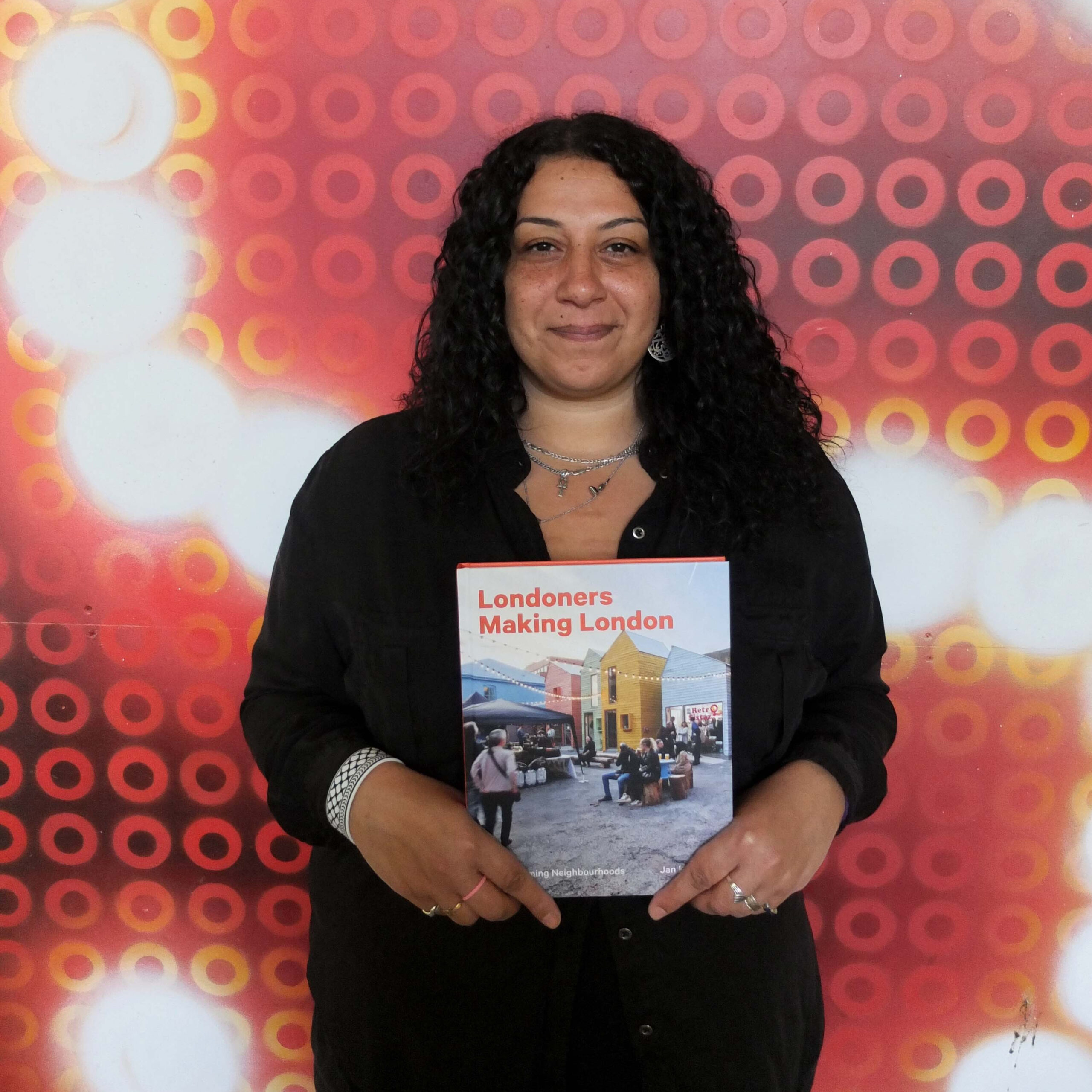 Photograph of Rasha holding the book she's featured in, smiling and looking at the camera, posing in front of a big graffiti 'S', outside Maxilla Men's Shed.